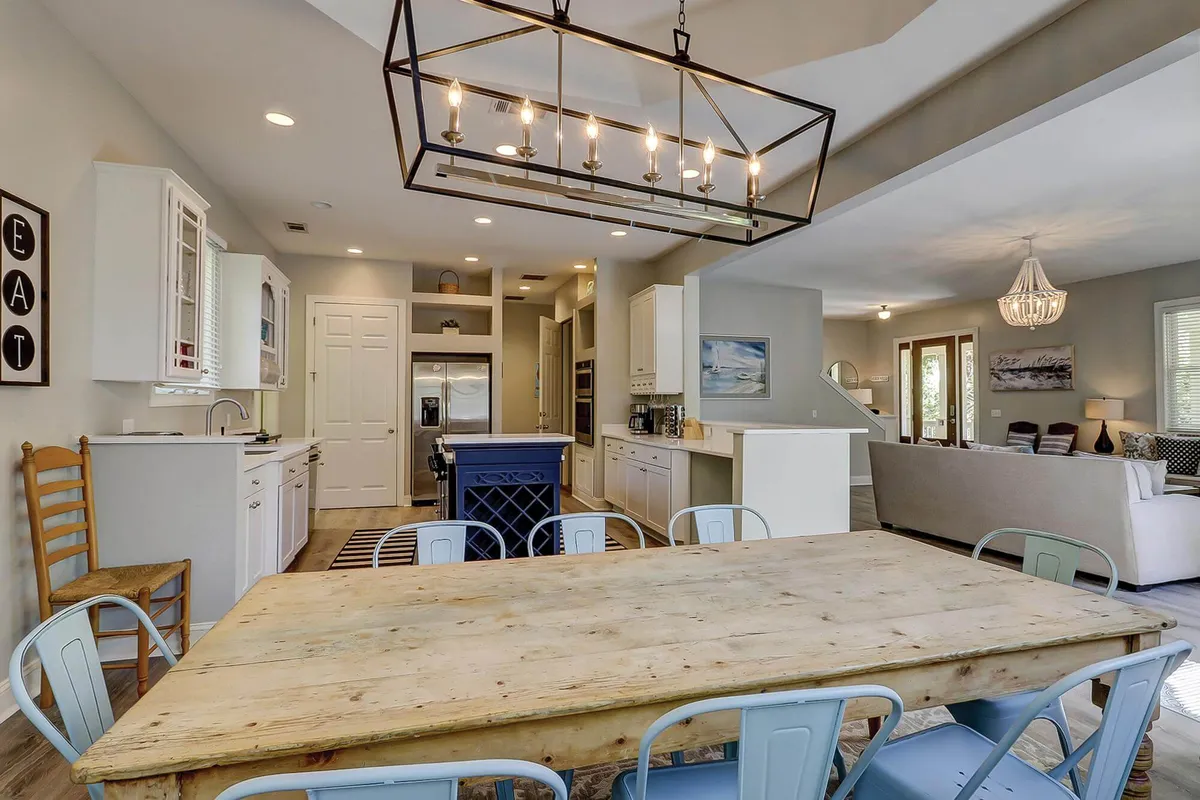 This image showcases a large wooden dining table set beneath a modern light fixture, seamlessly connecting to an open kitchen layout and a comfortable living area. The scene radiates warmth and togetherness, highlighting generous seating, natural light, and a unified space designed for gatherings.
