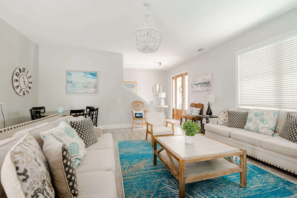 A spacious, inviting living room with plush sofas adorned with decorative pillows, a coffee table at the center, a large area rug, attractive wall art, and a prominent clock. The room is bathed in natural light streaming through generous windows, creating a welcoming atmosphere for family and friends to gather, relax, play games, or enjoy special occasions.