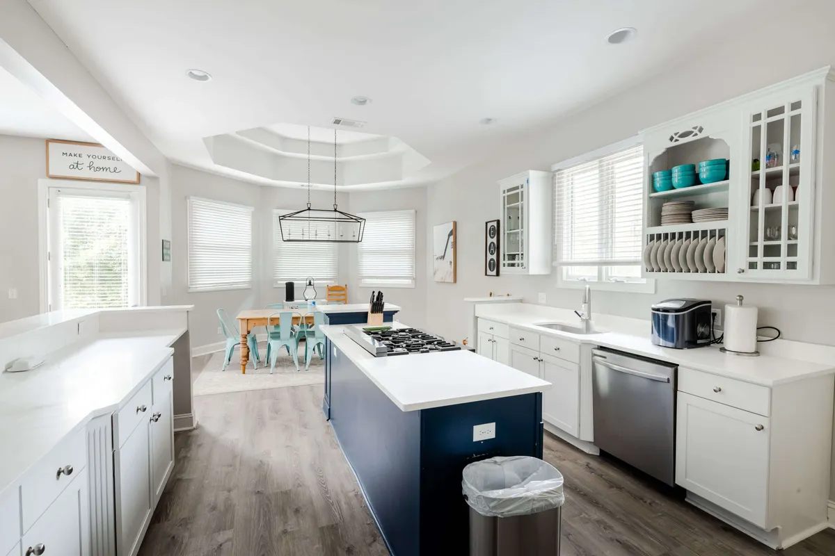 A view focused on the central kitchen island, adjacent dining area, and cabinetry with displayed dishware. The arrangement invites activities like baking, shared meals, and entertaining, while ample daylight streams through unseen windows, enhancing the welcoming mood.
