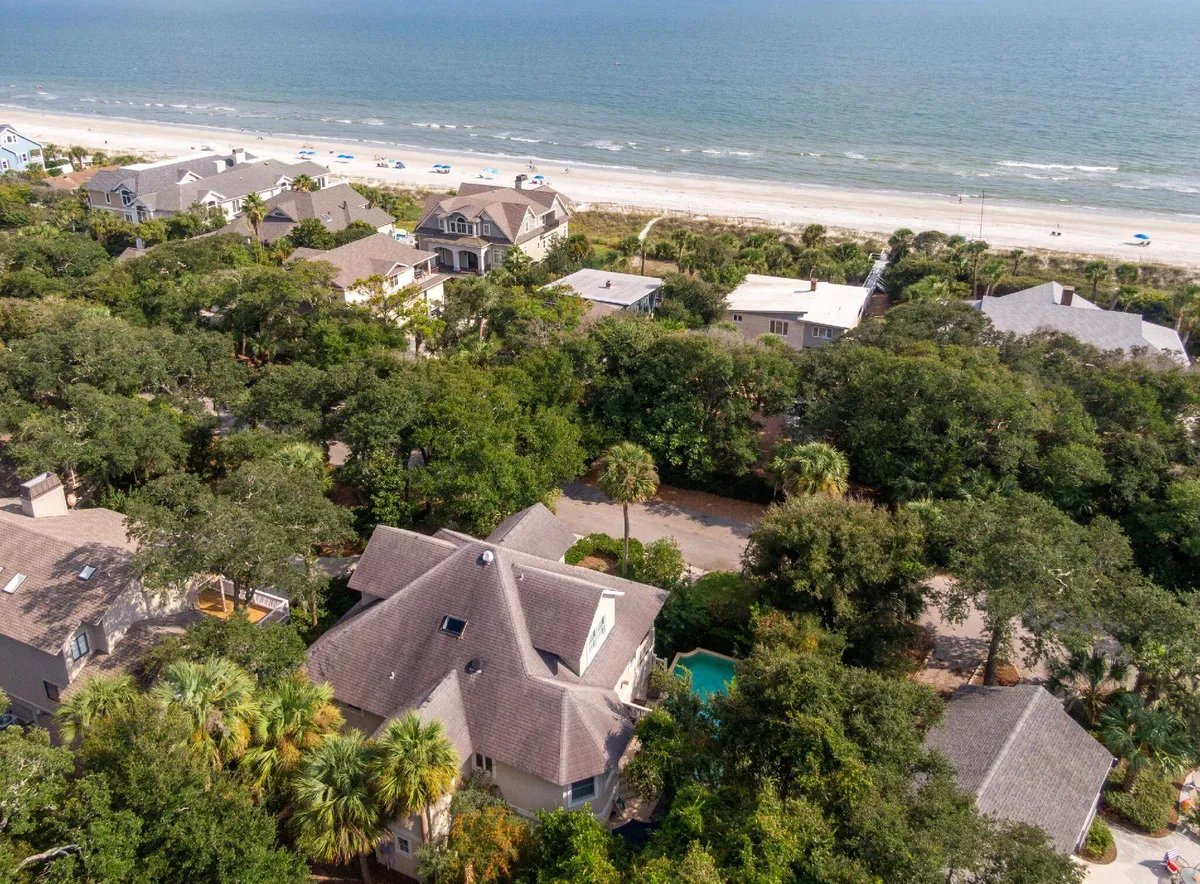 A panoramic view reveals the close arrangement of the property, its pool, and the nearby beach. The image captures the seamless transition between the yard, pool area, and the sandy shoreline, with lush greenery and neighboring houses completing the scene.