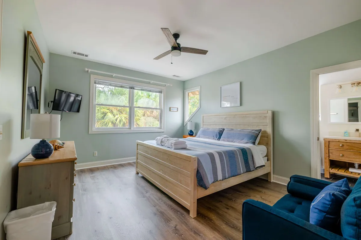 A tranquil bedroom featuring a comfortable bed adorned with plush bedding, a large window letting in natural light, a serene blue chair by the side, and a dresser with tasteful decor. The setting invites relaxation, reading, and restful sleep, with soft colors and calming design elements.