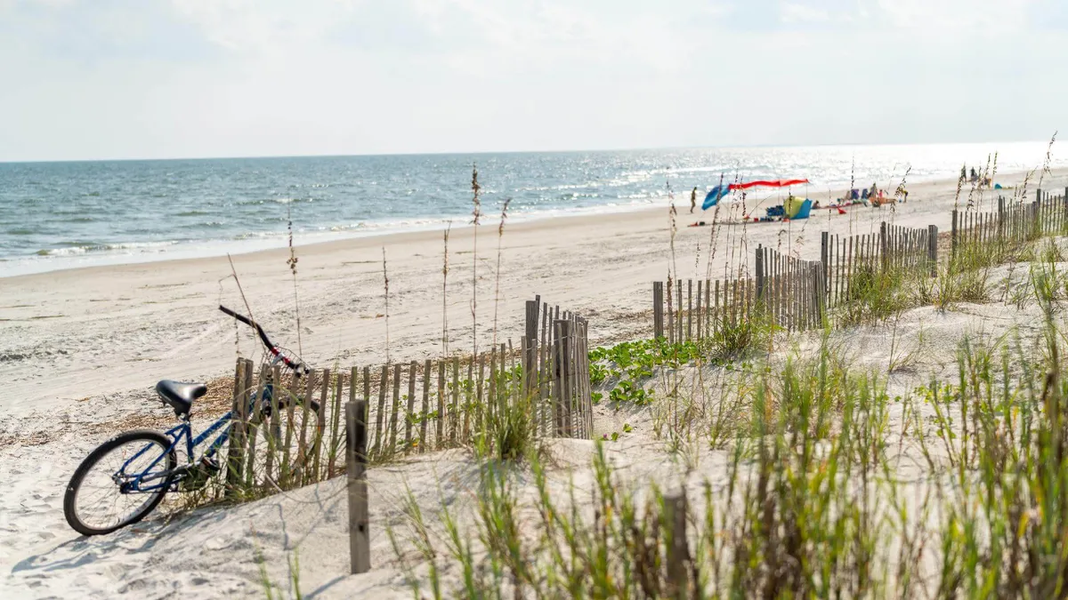 A sun-drenched stretch of beach with bicycles parked on the sand, flanked by dunes and umbrellas. The ocean glimmers in the background, evoking a sense of adventure and easy access to outdoor activities.