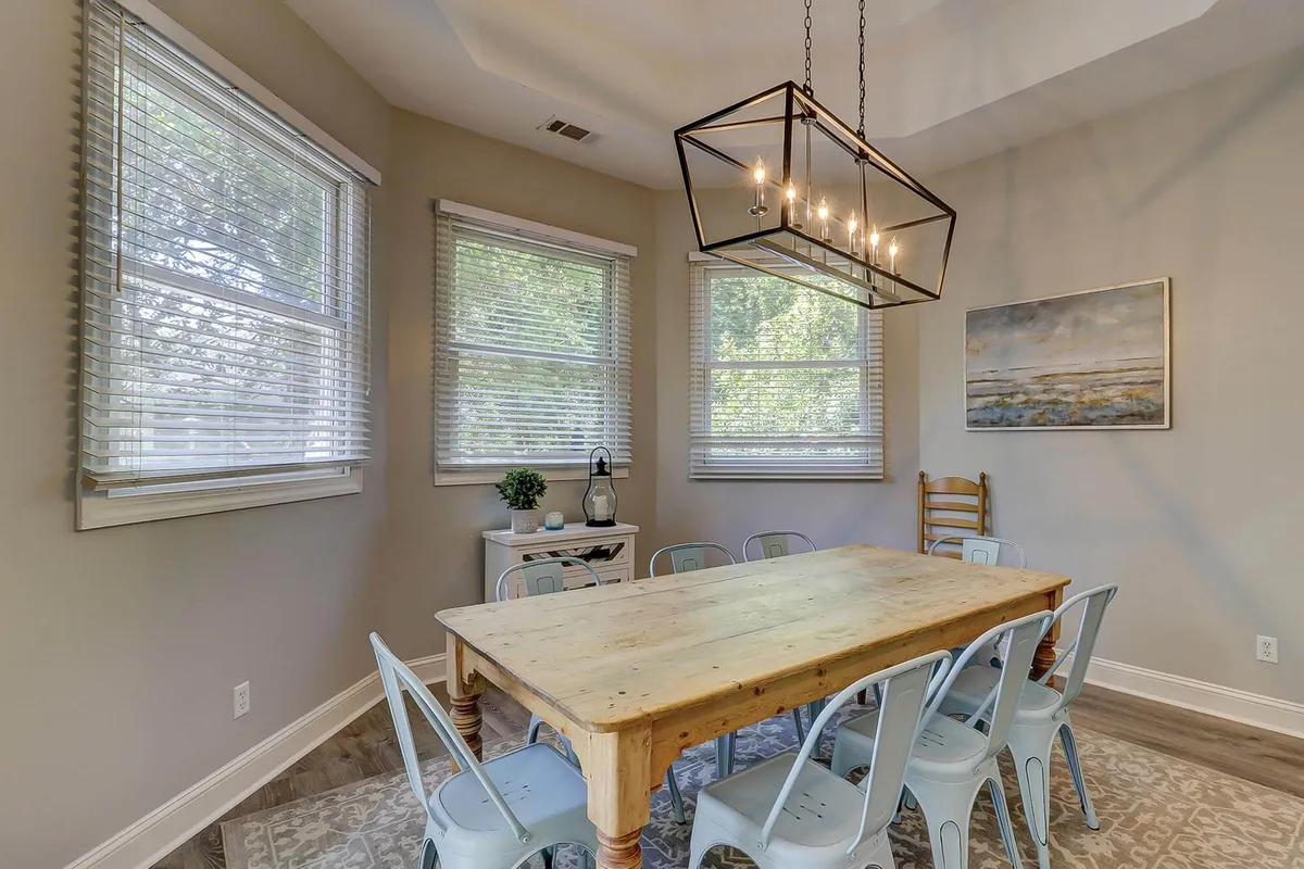An inviting dining area featuring a sturdy wooden table surrounded by stylish blue chairs, with a modern chandelier overhead and tasteful artwork on the wall. The space is clearly set up for group meals, celebrations, and intimate gatherings, offering a warm and welcoming ambiance.