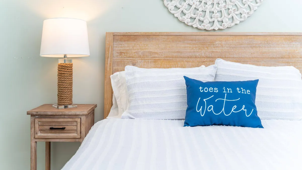 A bright, serene bedroom with a cozy bed adorned with decorative pillows, a bedside lamp, and a nightstand. The soft lighting and calming colors create an inviting space ideal for quiet moments such as reading or enjoying a morning coffee in bed.